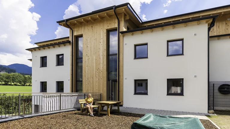 Person sitzt auf Holzbank und im Hintergrund ist die Wohnanlage Pfarrwerfen zu sehen